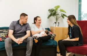 Women breastfeeding baby next to partner whilst speaking to a healthcare professional
