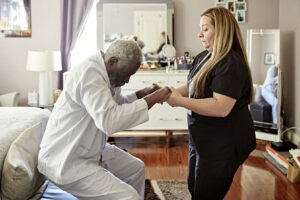 Side view of Black man in early 70s wearing pajamas and getting up with assistance of Latin American home caregiver in uniform.
