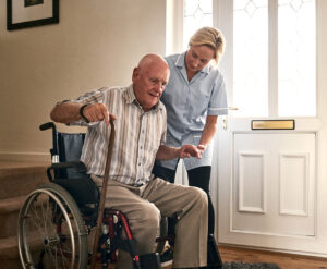 Female carer helping disabled senior man to get up from wheelchair at care home