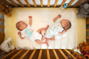 Two infants are lying side-by-side asleep in a crib. Stuffed animal toys surround the crib and a baby mobile hangs above. The infants are wearing dinosaur themed pajamas.