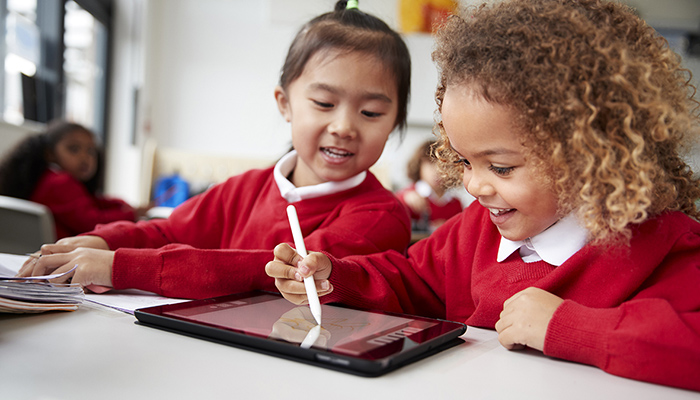 Close up of two kindergarten schoolgirls wearing school uniforms, sitting at a desk in a classroom using a tablet and stylus, looking at screen and smiling